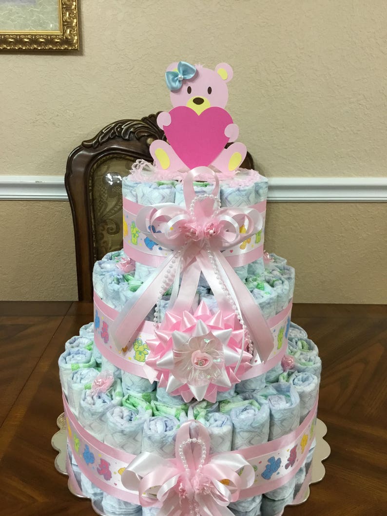 May include: A three-tiered nappy cake decorated with pink and white ribbons, a pink flower, and a pink bear holding a pink heart. The cake is made of rolled nappies and is a popular baby shower gift.