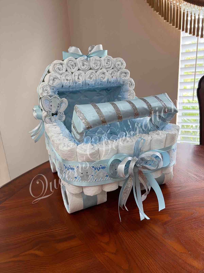 May include: A blue and white diaper cake shaped like a baby carriage. The carriage is decorated with a silver ribbon and a blue bow. The cake is made of diapers, rolled up and stacked. It is a baby shower gift.