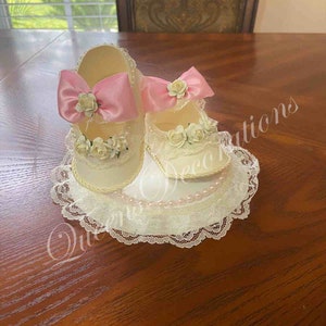 May include: Two white baby shoes with pink bows and white lace trim, sitting on a white lace doily with pearl accents.