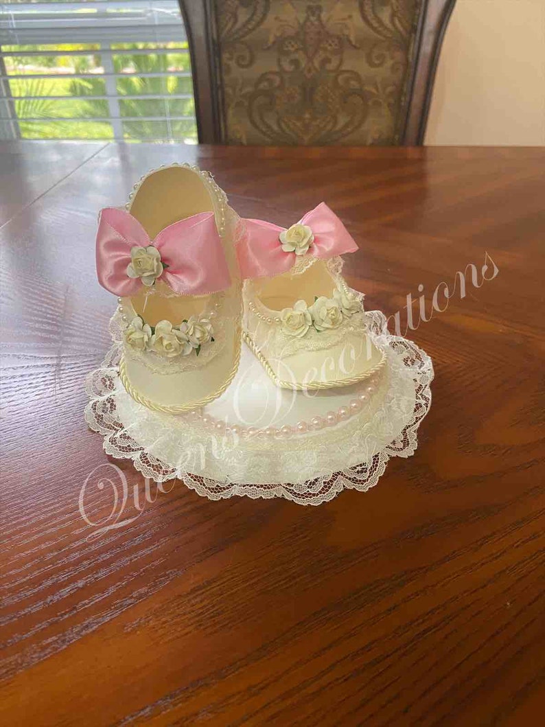 May include: Two white baby shoes with pink bows and white lace trim, sitting on a white lace doily with pearl accents.