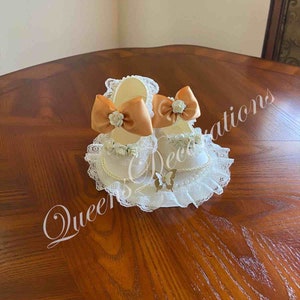May include: Two white satin baby shoes with lace trim and a peach-coloured satin bow with a white flower accent. The shoes are on a white lace base with a butterfly design.