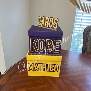 K&ouml;nnte beinhalten: Zwei &uuml;bereinander gestapelte Kartons f&uuml;r Karten, der obere Karton ist lila mit dem Wort "CARDS" in gelben Buchstaben, der untere Karton ist gelb mit den Worten "KOBE MATHIEU" in lila Buchstaben.