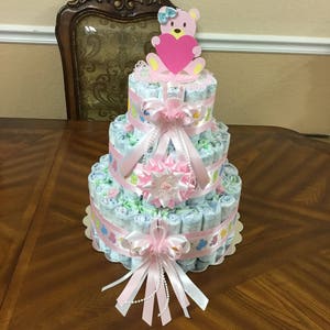 May include: A three-tiered nappy cake decorated with pink and white ribbons, a pink teddy bear holding a heart, and pearl beads. The cake is on a wooden table.