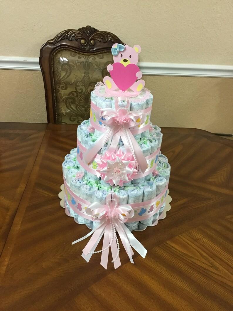 May include: A three-tiered nappy cake decorated with pink and white ribbons, a pink teddy bear holding a heart, and pearl beads. The cake is on a wooden table.