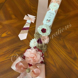 May include: A pink and gold sash with a white tag that says "Mommy To Be" and a large bow. The sash is decorated with burgundy, white, and gold flowers. A matching tie with a burgundy, white, and gold striped pattern is also included.
