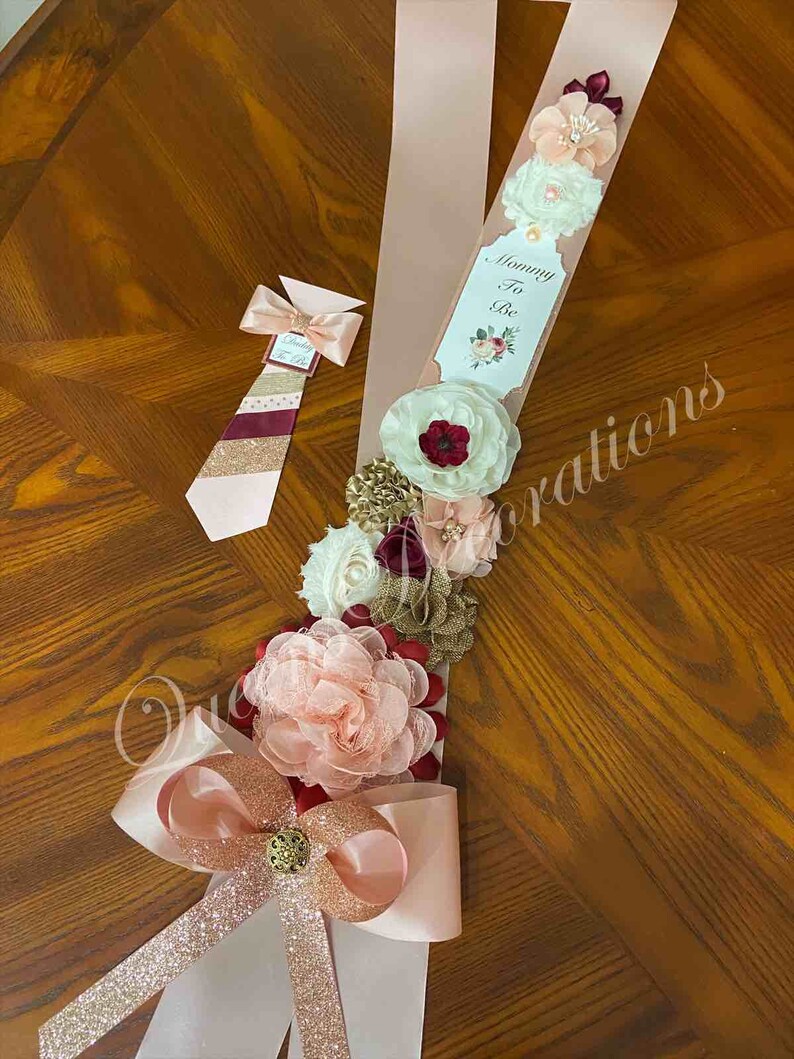 May include: A pink and gold sash with a white tag that says "Mommy To Be" and a large bow. The sash is decorated with burgundy, white, and gold flowers. A matching tie with a burgundy, white, and gold striped pattern is also included.