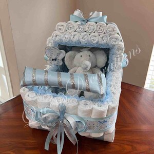 May include: A blue and white diaper cake with a stuffed elephant toy inside. The cake is decorated with blue and silver ribbon and a bow.