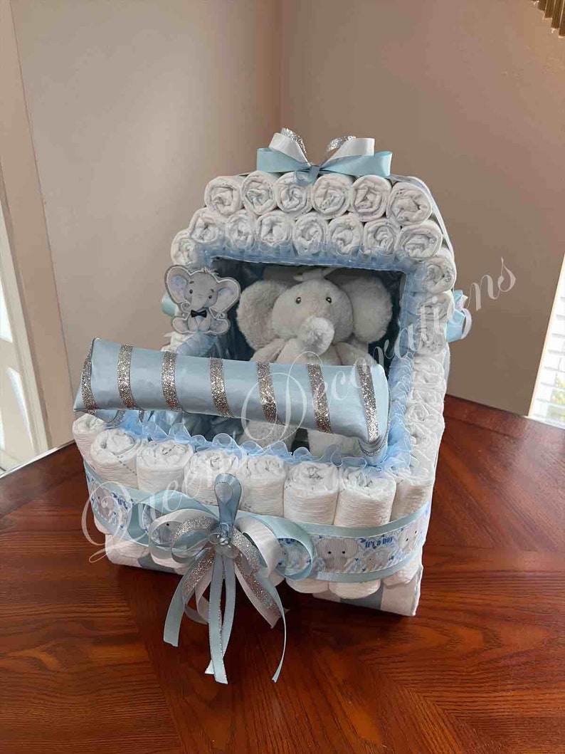 May include: A blue and white diaper cake with a stuffed elephant toy inside. The cake is decorated with blue and silver ribbon and a bow.