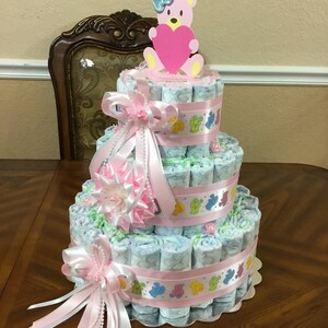 May include: A three-tiered nappy cake decorated with pink and white ribbons, a pink and white flower, and a pink teddy bear holding a pink heart. The cake is made of rolled up nappies and is decorated with a pink and white ribbon.