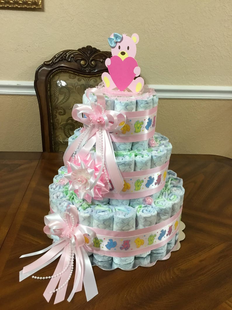 May include: A three-tiered nappy cake decorated with pink and white ribbons, a pink and white flower, and a pink teddy bear holding a pink heart. The cake is made of rolled up nappies and is decorated with a pink and white ribbon.