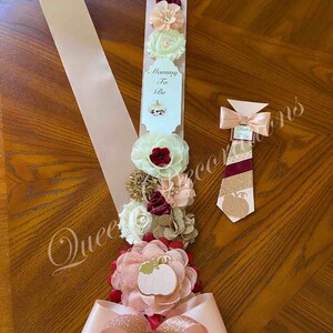 May include: A pink and gold satin sash with a bow and a pumpkin-shaped embellishment. The sash has a white tag that says "Mommy To Be". There are also several burgundy, peach, and white fabric flowers on the sash. A matching tie with a pumpkin design is also included.