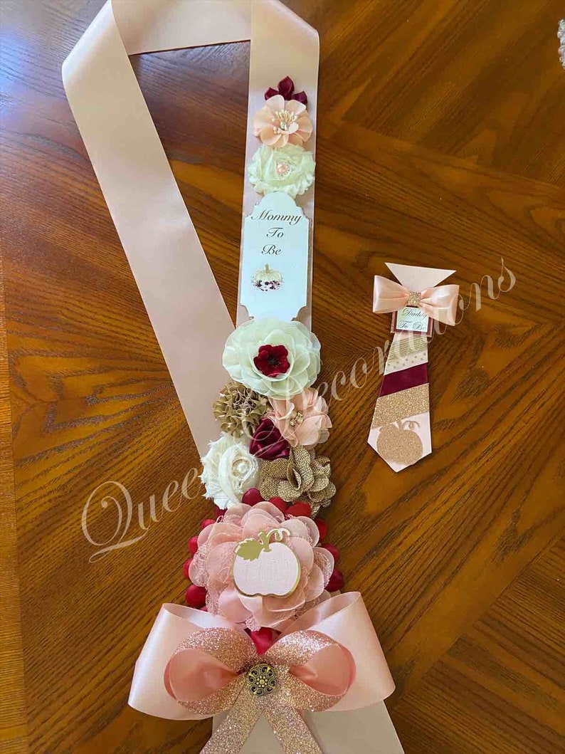 May include: A pink and gold satin sash with a bow and a pumpkin-shaped embellishment. The sash has a white tag that says "Mommy To Be". There are also several burgundy, peach, and white fabric flowers on the sash. A matching tie with a pumpkin design is also included.
