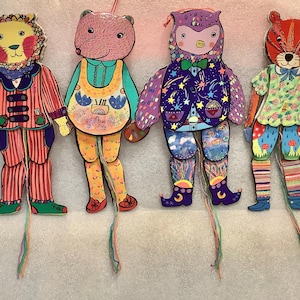 May include: Four colorful, whimsical paper dolls in the shape of animals: a lion, a bear, an owl, and a tiger. Each doll is dressed in unique, patterned clothing and has a string attached for hanging or play.