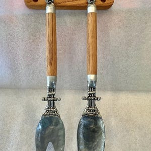 May include: A set of salad servers with wooden handles and ornate silver-toned metal details. The serving utensils are attached to a wooden hanging bar. The fork and spoon have hammered metal serving heads.