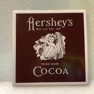 May include: A brown square tile with a white Hershey's Cocoa logo. The logo features a cherub holding a cup and sitting inside a cocoa pod. The text "Hershey's" is in a white cursive font with the text "REG. U.S. PAT. OFF." below it. The text "TRADE MARK" is below the cherub image and the word "COCOA" is in a large white font below the text "TRADE MARK".