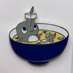 May include: A gray narwhal with a white belly and a white cube on its horn is swimming in a blue bowl of yellow soup with white cubes and green seaweed.