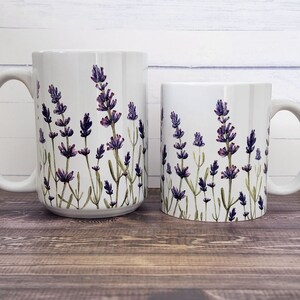 May include: Two white ceramic mugs with a lavender floral design. The larger mug is on the left, and the smaller mug is on the right. The lavender design features purple flowers and green stems. The mugs are set against a light wood background.