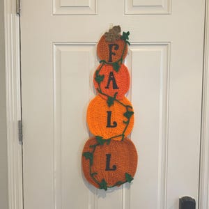 May include: A handmade crochet decoration for a door, featuring three stacked orange pumpkins spelling out "FALL" in black letters. Green vines and leaves accent the pumpkins, creating a festive autumn theme.