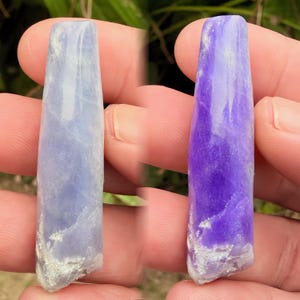 Puede incluir: Se muestran dos colgantes de piedras preciosas pulidas, uno azul claro y otro morado. Cada colgante tiene forma rectangular con una base ligeramente rugosa y texturizada. Las piedras preciosas se sostienen en una mano, mostrando su color y textura.