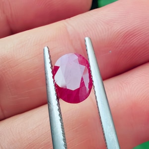 Puede incluir: Una gema de rubí facetada, de forma ovalada, sujeta con pinzas plateadas. El rubí muestra un color rojo intenso con facetas internas que reflejan la luz. Las pinzas son sujetadas por una mano, con el fondo desenfocado.