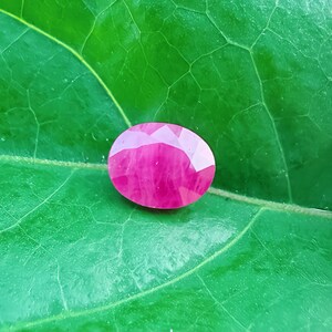 May include: A faceted, oval-shaped ruby gemstone, displaying a vibrant pinkish-red hue. The gem is set against a textured, green leaf background, highlighting its brilliance and colour. This gemstone is ideal for jewellery making.