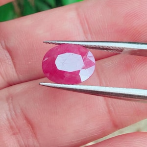 May include: A faceted, oval-shaped ruby gemstone. The ruby is a vibrant red color with a polished surface, held by a pair of silver tweezers. The gemstone's facets reflect light, creating a sparkling effect. The background is a blurred view of a hand.