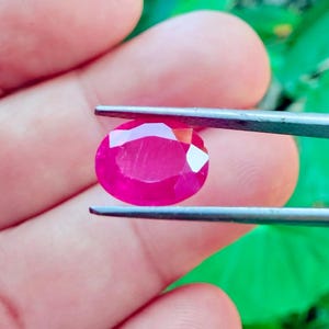 May include: A vibrant, oval-cut ruby gemstone held by tweezers. The ruby displays a deep, saturated red color with facets that catch the light. The background is a soft green, providing contrast to the gemstone.