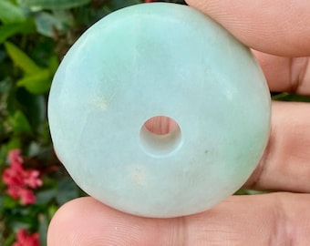 May include: A round, pale green jade pendant with a central hole. The stone has a smooth, polished surface and a subtle variation in color, with areas of lighter and darker green. The pendant is held in a person's hand.