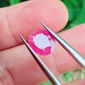 May include: A round, faceted ruby gemstone held by silver tweezers. The gemstone is a vibrant pinkish-red colour, with multiple facets reflecting light. The background is blurred, with hints of green.