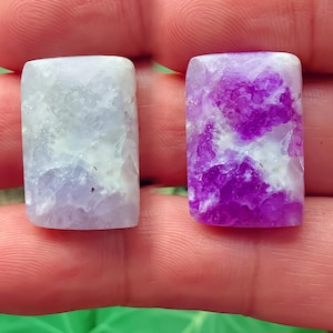 Puede incluir: Dos piedras preciosas rectangulares. Una es de color lavanda pálido con vetas blancas. La otra es de un púrpura vibrante con veteado blanco. Ambas piedras tienen una superficie pulida y lisa y se sostienen en una mano.