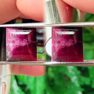 May include: Two rectangular, faceted gemstones with a deep magenta hue and internal white veining are held by tweezers. The gems have a polished, reflective surface, showcasing their clarity and colour. The background is blurred, with hints of green.