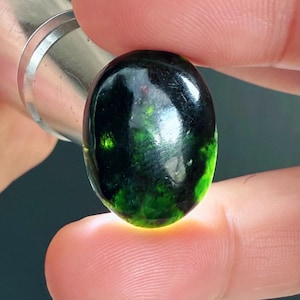 May include: A dark green, oval gemstone with a polished surface, held between fingers. The stone's colour varies from deep green to black. A metallic object is visible in the background. The gem reflects light, highlighting its glossy finish.