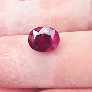 May include: A faceted, oval-shaped ruby gemstone. The ruby is a deep, rich red color with multiple facets that reflect light. The gemstone is held in a person's hand, with the skin tone appearing light pink.