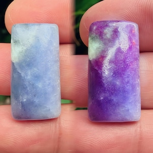 Puede incluir: Dos piedras preciosas rectangulares, una azul claro y otra morada, se sostienen entre los dedos. La piedra azul tiene vetas blancas y grises, mientras que la piedra morada tiene vetas blancas y rojas. Ambas piedras tienen una superficie pulida y lisa.