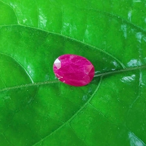 Pode incluir: Uma gema de rubi facetada, em forma oval, exibindo um tom magenta vibrante. A gema está sobre um fundo de uma folha verde-escura brilhante, criando um contraste marcante. O corte do rubi permite que a luz se reflita, mostrando seu brilho.