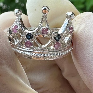 Puede incluir: Un anillo de corona plateado con piedras preciosas rosas y azul oscuro. El anillo, sostenido por una mano enguantada, presenta un recorte en forma de corazón y una banda decorativa. El diseño de la corona incorpora pequeñas piedras preciosas redondeadas.