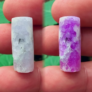 Puede incluir: Se muestran dos pequeños cilindros rectangulares de piedras preciosas. Uno es de color lavanda pálido y translúcido, y el otro es de color púrpura vibrante con vetas blancas. Ambos tienen una superficie rugosa y texturizada y una pequeña inclusión negra.