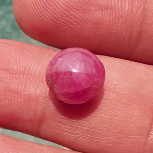 Puede incluir: Primer plano de una gema rosa, redonda y facetada. La gema se sostiene en la mano de una persona, con el tono de la piel como fondo contrastante. La superficie de la gema refleja la luz, mostrando sus facetas pulidas y su color vibrante.