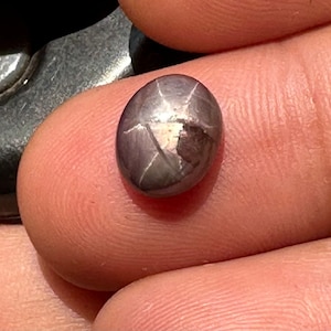 May include: A dark gray star-shaped gemstone with a faceted cut. The gemstone is oval-shaped and has a shiny, reflective surface.