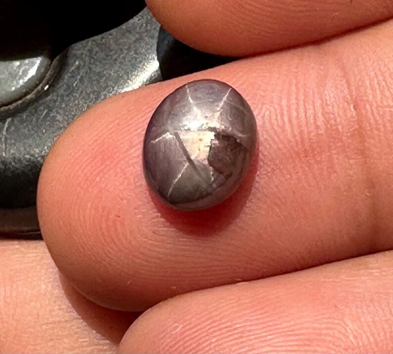 May include: A dark gray star-shaped gemstone with a faceted cut. The gemstone is oval-shaped and has a shiny, reflective surface.