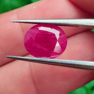 Könnte beinhalten: Ein leuchtender, oval geschliffener Rubin mit tiefem, pink-rotem Farbton und facettierter Oberfläche. Der Edelstein wird zwischen den Spitzen einer Metallpinzette gehalten und zeigt seine durchscheinende Qualität sowie polierte Facetten.