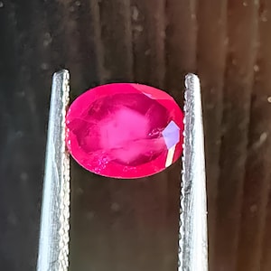 May include: A vibrant, oval-cut ruby gemstone held by tweezers. The ruby displays a deep red hue with facets catching the light, showcasing its brilliance. The gemstone is the focal point, suggesting jewelry or gemstone sales.