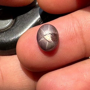 May include: A dark gray oval-shaped gemstone with a star-shaped pattern in the center. The gemstone is set in a silver setting.