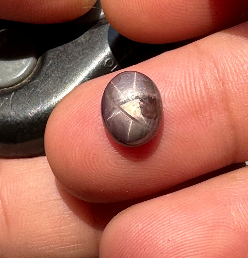 May include: A dark gray oval-shaped gemstone with a star-shaped pattern in the center. The gemstone is set in a silver setting.