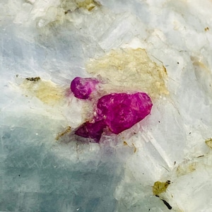 Pode incluir: Close-up de uma gema de rubi rosa incrustada em uma rocha branca. O rubi é cercado por cristais brancos e cinzentos.