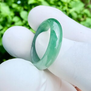 May include: A smooth, polished ring crafted from translucent green jadeite. The band features a rounded profile with a glossy finish, showcasing natural variations in color intensity ranging from pale mint to deep emerald green throughout the stone.