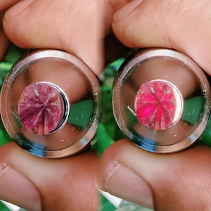 May include: Two close-up views of a metallic object with a circular, polished metal rim. The centre of each object features a vibrant, reddish-pink gemstone with a radial pattern. The objects are held in a person's hands.