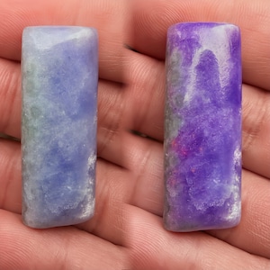 Puede incluir: Dos piedras preciosas rectangulares, una en tonos azules y verdes, la otra en morado y rosa. Ambas tienen una superficie pulida y una parte superior ligeramente redondeada. Las piedras se sostienen en una mano, mostrando sus colores y texturas naturales.