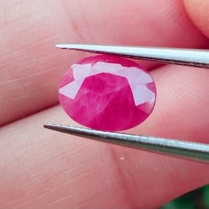 Puede incluir: Una gema de rubí facetada, de forma ovalada, sujeta con pinzas. La gema es de un color rosa-rojo vibrante, con múltiples facetas que reflejan la luz. El fondo está borroso, resaltando los detalles de la gema. Esta imagen muestra una piedra preciosa.