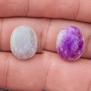 May include: Two oval-shaped gemstones. One is a pale, milky white, and the other is a vibrant purple with white marbling. Both are smooth and polished, showcasing natural patterns. The gemstones are held in a hand.